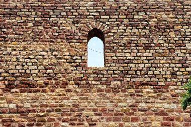 Kırsal taştan bir duvar, açık mavi gökyüzünü çerçeveleyen dar kemerli bir pencereye sahiptir. Yıpranmış duvarcılık, sıcak toprak tonları ve dokulu yüzeyler tarihi cazibeyi ve antik mimari ayrıntıları vurgular..