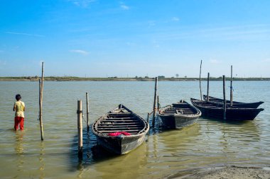 Tahta tekneler, rıhtımlar ve sığ sularda yüzen bir çocukla sakin sahil manzarası.