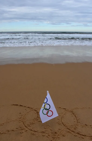 Anéis olímpicos desenhados na areia com uma bandeira olímpica . — Fotografia de Stock