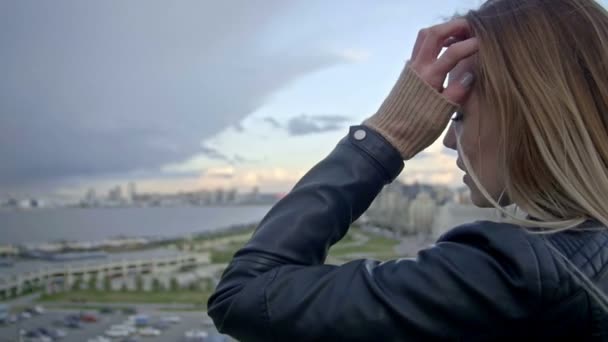 Ragazza carina con lunghi capelli biondi in giacca di pelle raddrizza i capelli guardando alla città moderna slo-mo — Video Stock