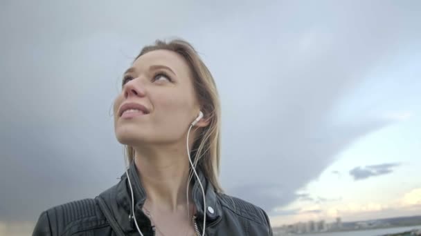 Roztomilá dívka s dlouhými blonďatými vlasy, v kožené bundě narovnává vlasy při pohledu na oblohu slo-mo — Stock video