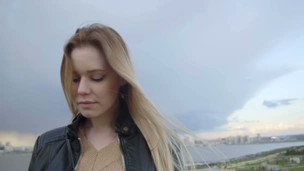 Roztomilá dívka s dlouhými blonďatými vlasy s modrým yeys v kožené bundě narovnává vlasy při pohledu na oblohu slo-mo — Stock video