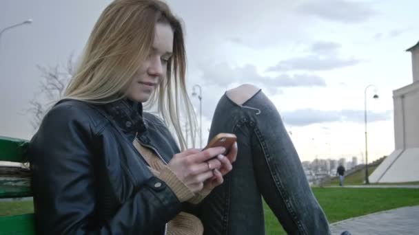 Söt glad flicka med långa blonda hår i skinnjacka rätar hår användning gadget sitter på bänken närbild — Stockvideo