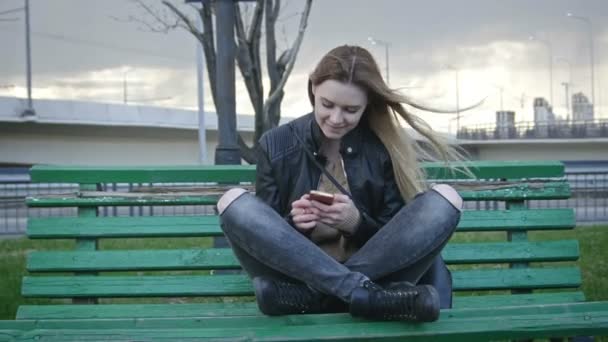 Schattig gelukkig meisje met lang blond haar in lederen jas rechtzetten haar gebruik gadget zittend op het bankje in de wind 4k — Stockvideo