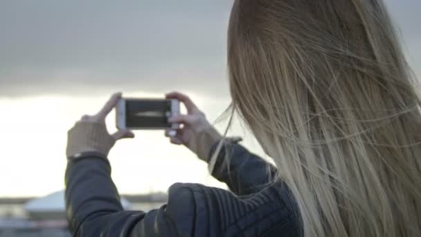 Ładna dziewczyna z długie blond włosy w skórzanej kurtce prostuje włosy fotografowanie miasta przez telefon — Wideo stockowe