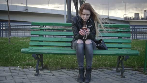 Разочарованная девушка с длинными светлыми волосами в кожаной куртке выпрямляет волосы с помощью гаджета сидит на скамейке на ветру слушать наушники широкий угол фронта — стоковое видео