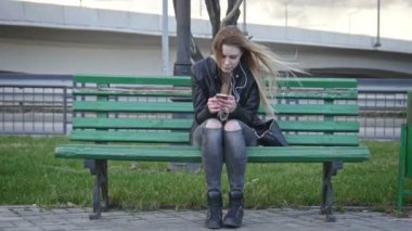 Deri ceketli uzun sarı saçlı dissapointed kız saç kullanım gadget Rüzgar dinle kulaklık 4 k geniş açı öndeki bankta oturmuş düzeltiyor