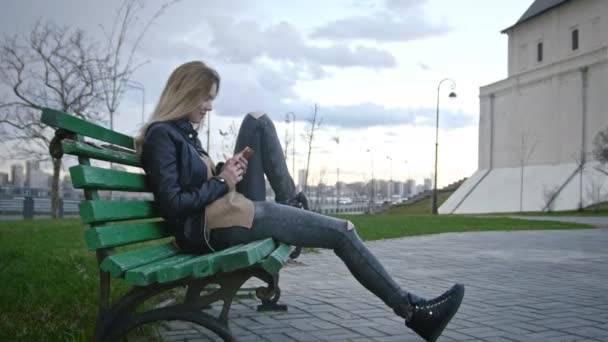Милая счастливая девушка с длинными светлыми волосами в кожаной куртке выпрямляет волосы с помощью гаджета сидит на скамейке — стоковое видео