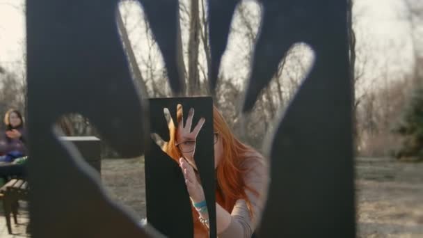 Belle fille aux cheveux roux portant des lunettes dans le parc du printemps regarde à travers monument abstrait — Video
