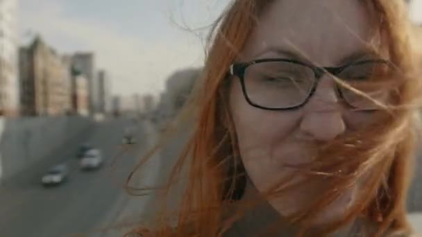 Красивая девушка с рыжими волосами в очках на улице возле шоссе смотрит в камеру — стоковое видео