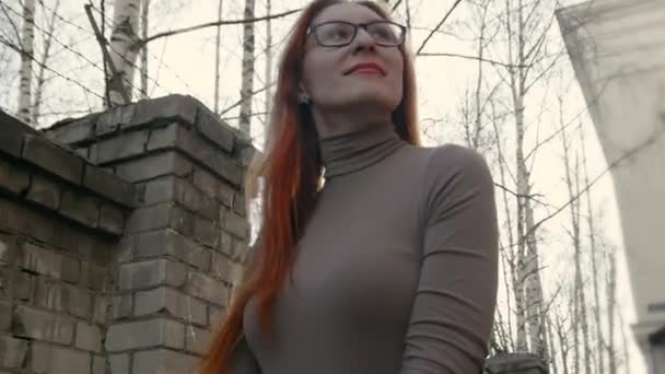 Belle fille aux cheveux roux portant des lunettes debout près du mur de briques et regarder à la caméra — Video