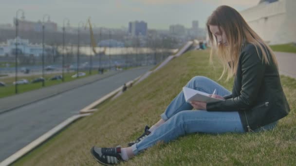 Linda chica joven escribiendo diario se sienta en un fondo de vistas de la ciudad al atardecer — Vídeo de stock