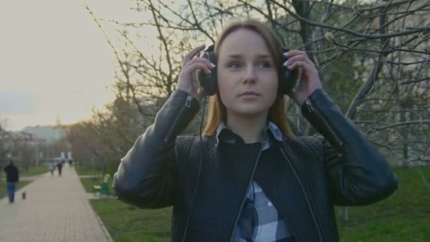 Linda chica joven se pone los auriculares en el parque al atardecer — Vídeos de Stock