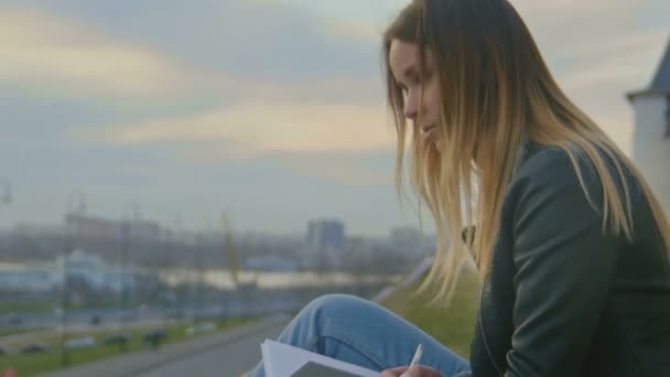 Милая молодая девушка, пишущая дневник, сидит на фоне городских пейзажей в сумерках крупным планом — стоковое видео