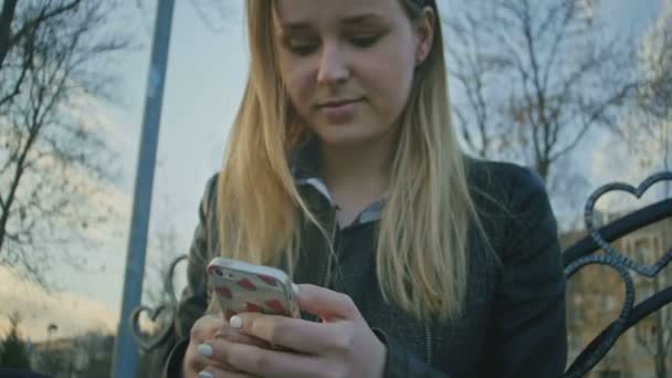 Linda chica joven utiliza el teléfono sentado en el banco al atardecer — Vídeo de stock