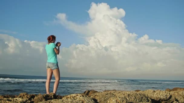 Junge Frau mit roten Haaren in Brille fotografiert Meer, das auf den Felsen vor Wolken steht, Strand der Dominikanischen Republik, Weitwinkel — Stockvideo