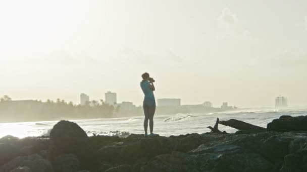 Bellezza giovane donna con i capelli rossi in occhiali va sulle rocce e fotografa la vita marina sul mare dei caraibi di fronte alle onde in Repubblica Dominicana, teleobiettivo, slow-motion — Video Stock