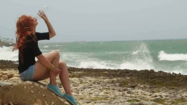 Baştan çıkarıcı kız içki içme suyu plastik şişe, fırtına denize sıfır plaj üzerinde oturan uzun kızıl saçlı yukarıdan aşağıya doğru yavaş hareket