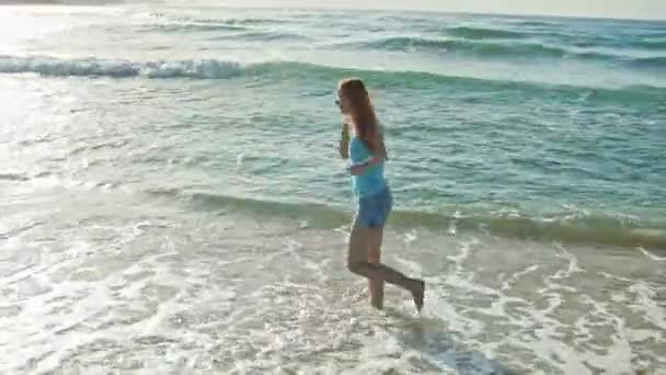 Mujer joven con el pelo largo y rojo en pantalones cortos juega con las olas corriendo, sintiendo el mar, el paisaje marino, la playa de República Dominicana — Vídeos de Stock