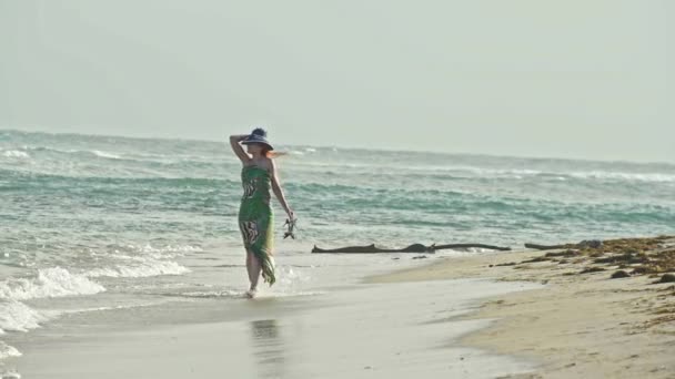 Jonge vrouw In lange jurk houden een hoed en wandelen langs de zee, slow-motion — Stockvideo