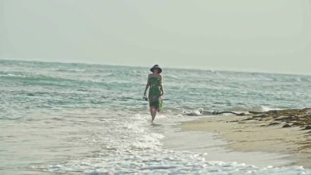 Jonge vrouw In lange jurk houden een hoed en wandelen langs de zee bij de waterkant, slow-motion — Stockvideo