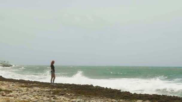 Attrayant jeune fille avec de longs cheveux roux debout sur la plage près de la mer de tempête, au ralenti — Video