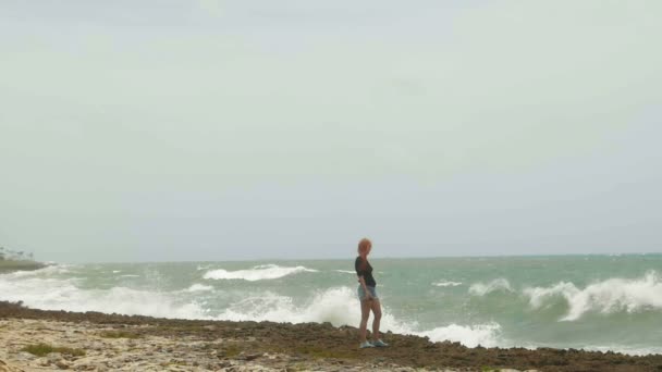 Aantrekkelijke jonge vrouw met lange rode haren permanent aan de kust en op zoek naar de storm-zee, vertraagd, groothoek — Stockvideo