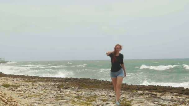 Mujer atractiva joven con el pelo rojo largo vestido en pantalones cortos caminando en la costa frente al mar de tormenta, gran ángulo, cámara lenta — Vídeos de Stock