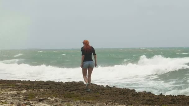 Jonge aantrekkelijke vrouw met lange rode haren gekleed in korte broek lopen aan de kust tegenover de bestorming zee — Stockvideo