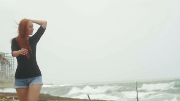 Giovane donna attraente con lunghi capelli rossi vestiti in pantaloncini corti a piedi lungo il bordo del mare tempesta, slow-motion — Video Stock