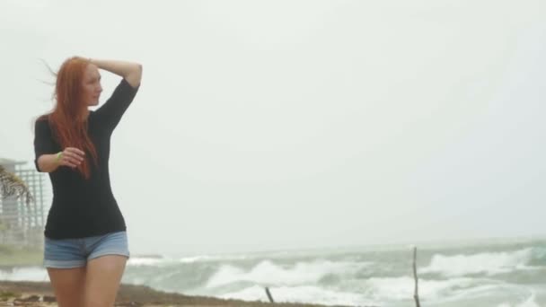 Mujer atractiva joven con el pelo rojo largo vestido en pantalones cortos caminando a lo largo del borde del mar de tormenta, cámara lenta — Vídeos de Stock