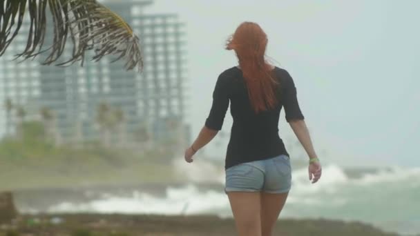 Jeune femme séduisante aux longs cheveux roux habillée en short court marchant sur la côte face à la mer orageuse, s'éloigner, ralentir — Video