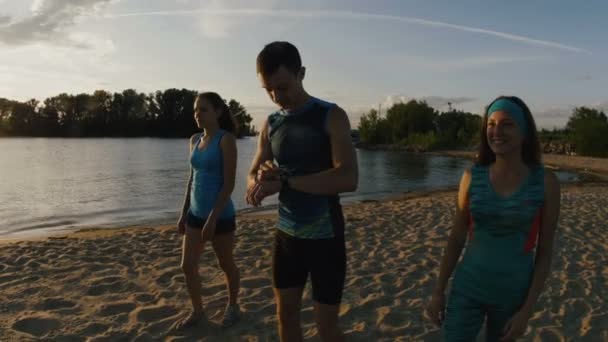 Um grupo de atletas - duas meninas e um cara corre na praia, perto do rio ao pôr do sol, usar gadget para medir a frequência cardíaca — Vídeo de Stock