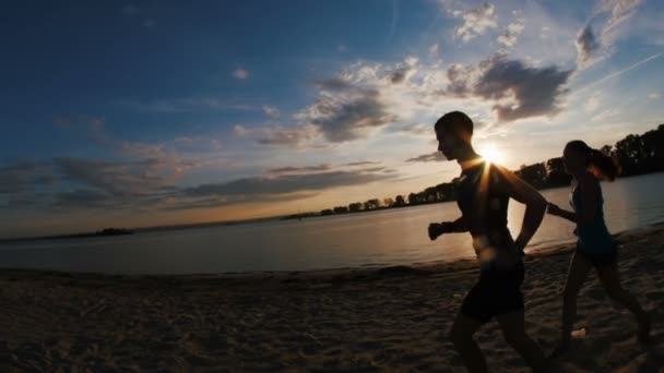 Група спортсменів - дві дівчини і хлопець біжить на пляжі, біля річки на заході сонця, силует, повільний рух, біжить повз — стокове відео