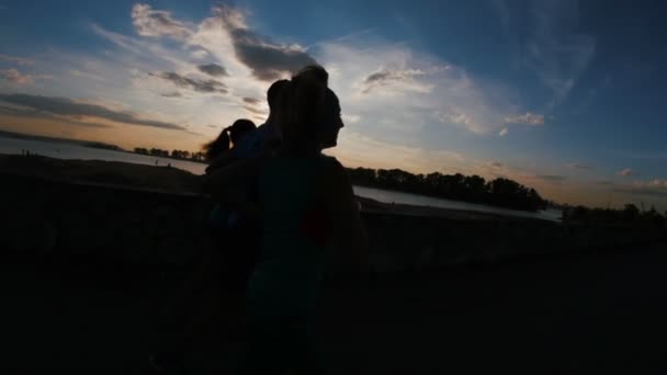 Un grupo de atletas - dos chicas y un tipo corriendo en el parque de la ciudad, cerca del río al atardecer, silueta, cámara lenta — Vídeo de stock