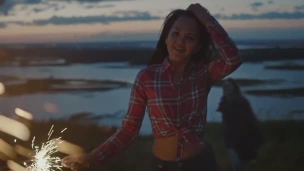 Sorrindo jovem dançando em uma colina alta com sparkler ao pôr do sol em câmera lenta — Vídeo de Stock