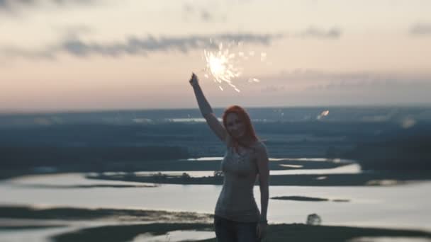 Lächelnde junge Frau mit Wunderkerze in der Abenddämmerung auf einem hohen Hügel — Stockvideo