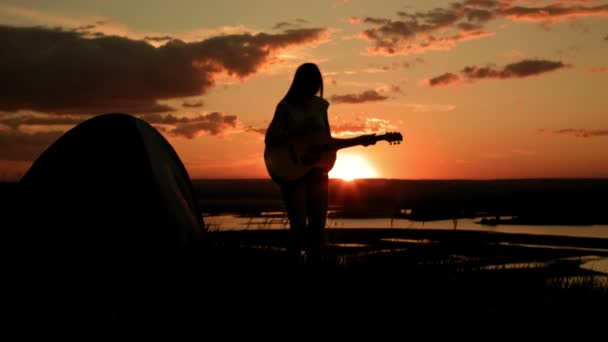 Atrakcyjna dziewczyna romantyczna gra na gitarze akustycznej, o zachodzie słońca w pobliżu camping na wysokim wzgórzu, sylwetka — Wideo stockowe