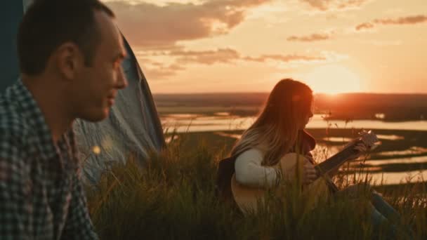 Menina atraente nova com amigos toca guitarra acústica no acampamento ao ar livre em alta colina no pôr do sol de verão — Vídeo de Stock