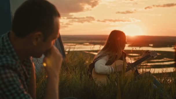 Junge attraktive Frau mit Freunden stimmt Akustikgitarre im Zelten im Freien auf einem hohen Hügel bei sommerlichem Sonnenuntergang — Stockvideo