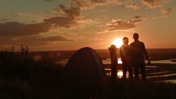 Familia joven - padre, madre e hijo en el camping - se encuentra en la colina alta al atardecer de verano y tienen feliz, silueta — Vídeo de stock