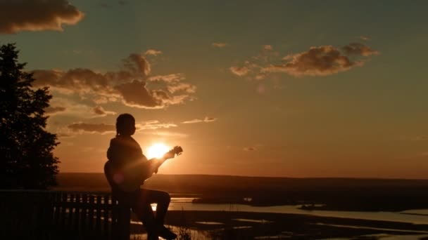 Jovem romântica tocando guitarra acústica, ao pôr do sol perto de acampar em alta colina, silhueta, ângulo largo — Vídeo de Stock