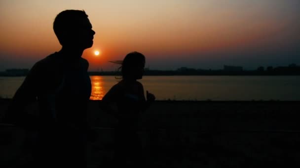 Pár fiatal felnőtt sportolók: nő és férfi futó sétány-folyó mentén a naplemente, silhouette, lassú mozgás — Stock videók
