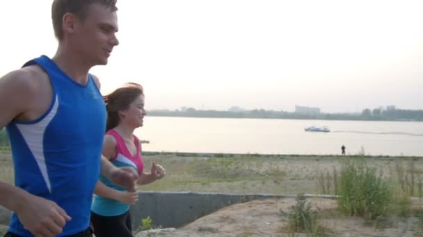Пара молодых взрослых спортсменов: женщина и мужчина бегут вдоль набережной реки. Концепция здорового образа жизни, замедленная съемка — стоковое видео
