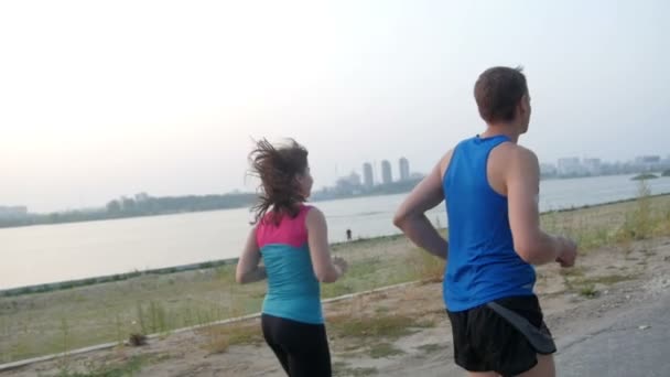 Пара молодых взрослых спортсменов: женщина и мужчина бегают по набережной реки в современном городе. Концепция здорового образа жизни, замедленная съемка, вид сзади — стоковое видео