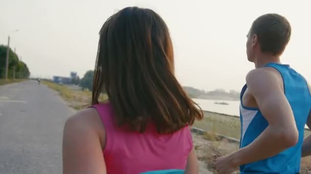 Paar van jonge volwassen atleten: vrouw en man loopt langs de promenade van de rivier bij zonsondergang. Gezonde levensstijl concept, achteraanzicht — Stockvideo