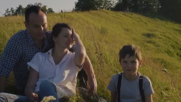 Familia feliz - padre, madre e hijo - sentado en lo alto de la colina al atardecer. Vacaciones de verano. mirando a la cámara — Vídeo de stock