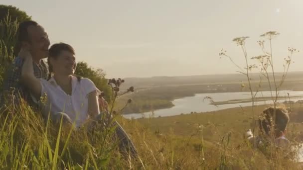 Glückliche Familie - Vater, Mutter und Sohn - lacht, sitzt auf einem hohen Hügel, in der Nähe des Sees bei Sonnenuntergang. Sommerferien — Stockvideo