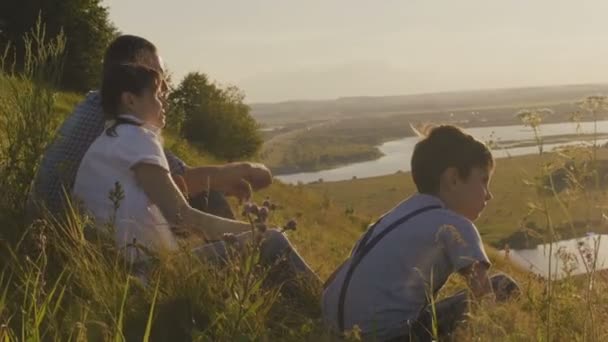 Lycklig familj - far, mor och son - sitter på hög kulle, nära sjön vid solnedgången. Sommar semester — Stockvideo