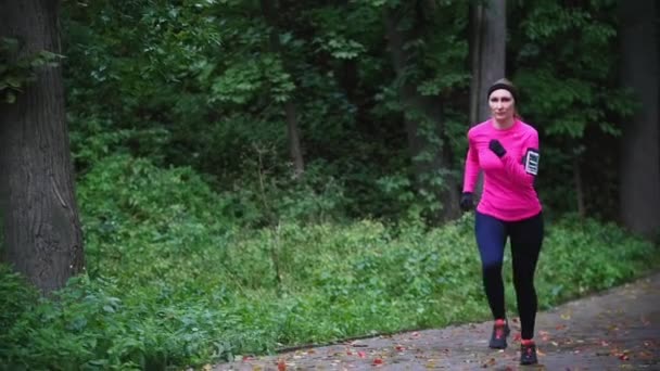 Corredor mujer joven corriendo en el parque de otoño haciendo ejercicio al aire libre en el clima sombrío lluvia, traje de entrenamiento rosa brillante, cámara lenta, primer plano — Vídeos de Stock